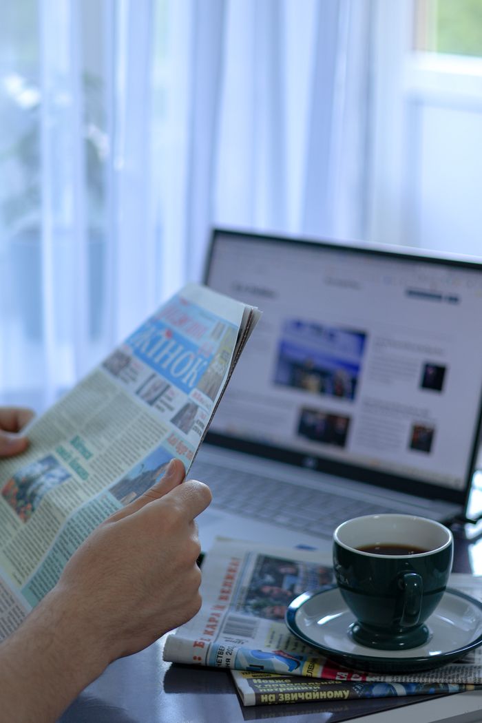 A person reads a newspaper while drinking coffee beside a laptop indoors.
